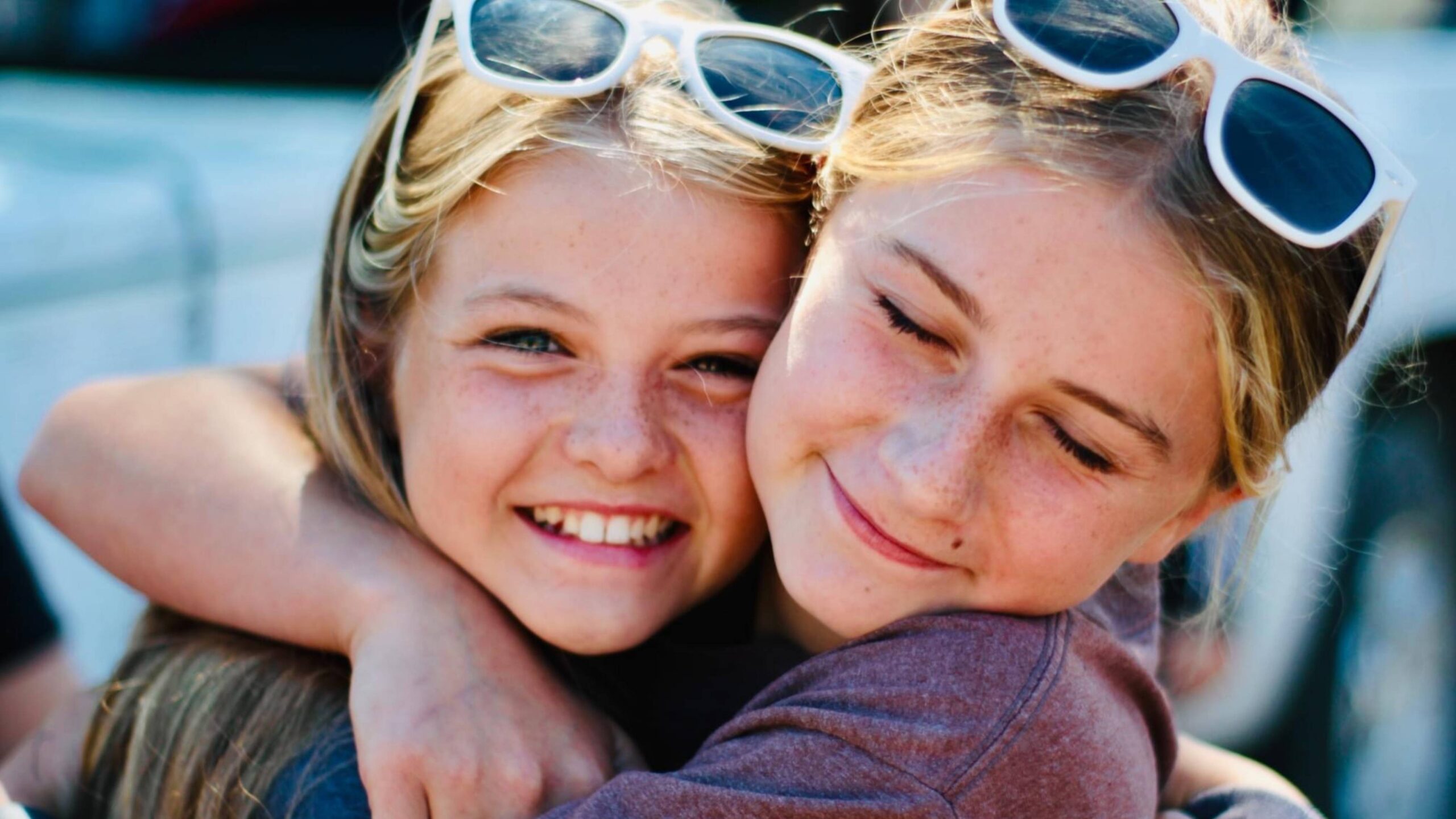 Two young girls wearing sunglasses, embracing warmly.