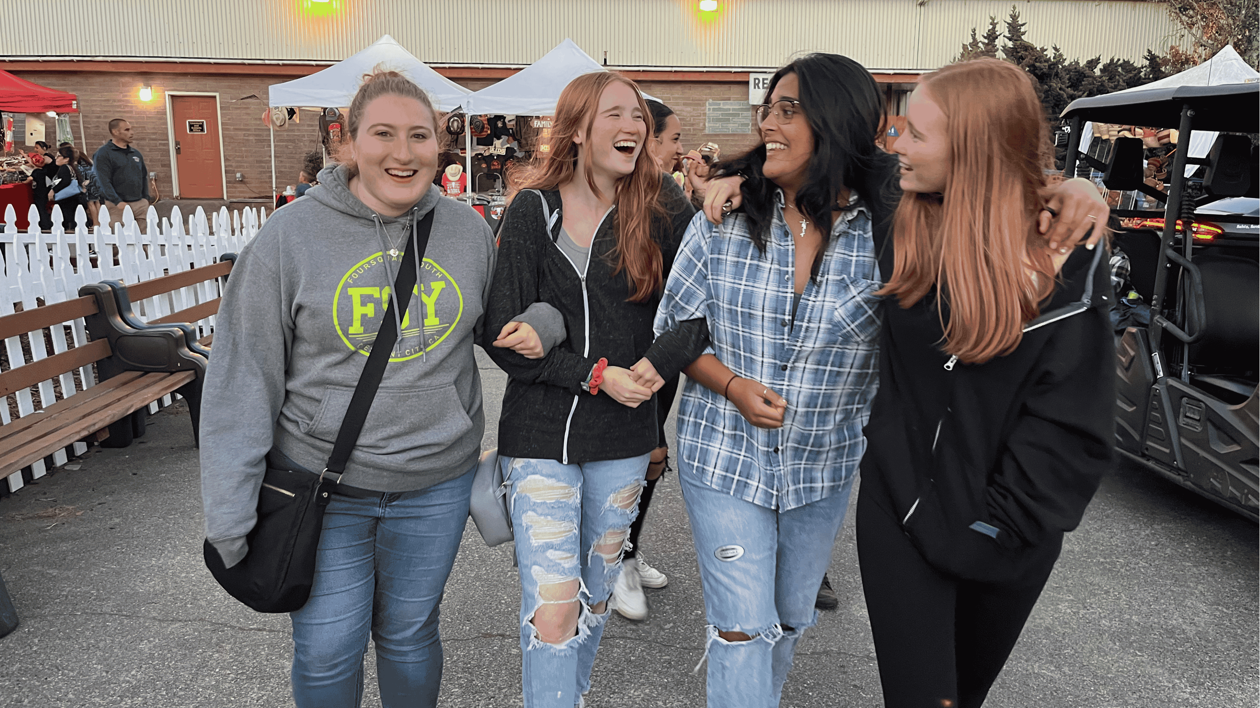 Four young women, laughing and walking together, are outside at an event, wearing casual clothing.
